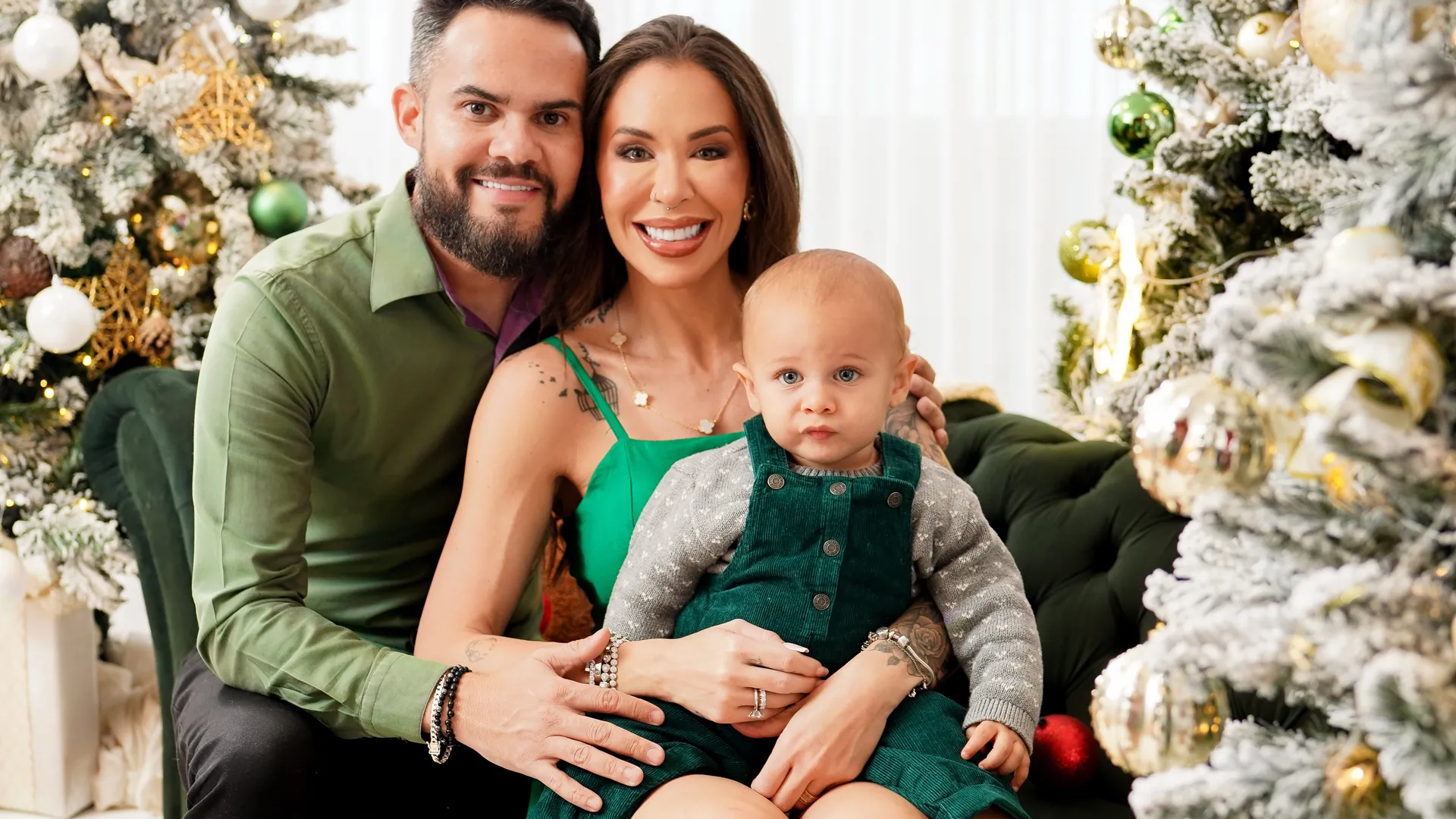 Pai, mãe e bebê num sofá de veludo verde entre árvores de Natal nevadas, ensaio de Natal em família no estúdio do Lago Sul, Brasília